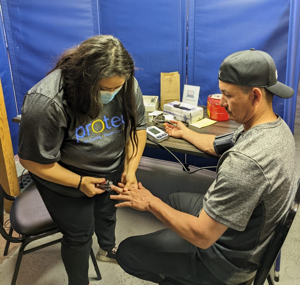 Agricultural health program at Proteus, Inc. Health worker places pulse oximeter on a farmworkers left hand while his right arm, wrapped in a blood pressure cuff, is resting on the table beside them. 